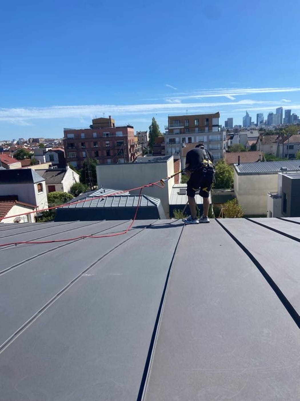 Couvreur harnaché et relié à une ligne de vie sur une toiture en zinc à joint debout, La Défense en arrière-plan.